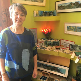 Picture of Annie in front of a display of her artwork and handmade books at her open house show at Sky Blue House in May 2019.